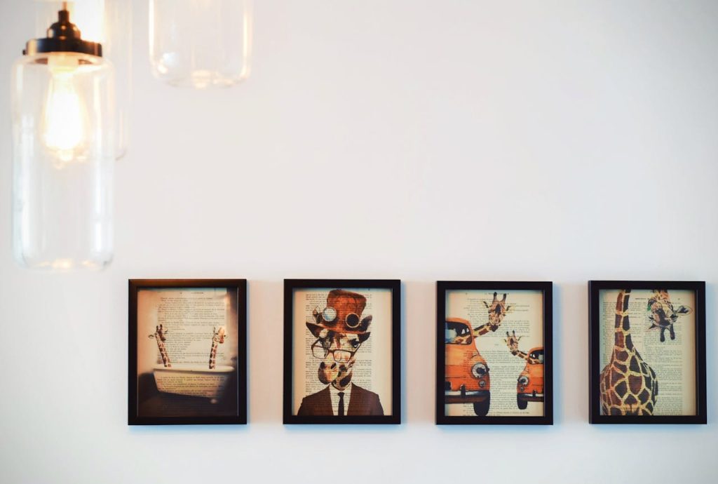 Four framed artworks featuring whimsical giraffe illustrations overlaid on vintage book pages, arranged in a row on a white wall, with hanging glass pendant lights partially visible in the upper left