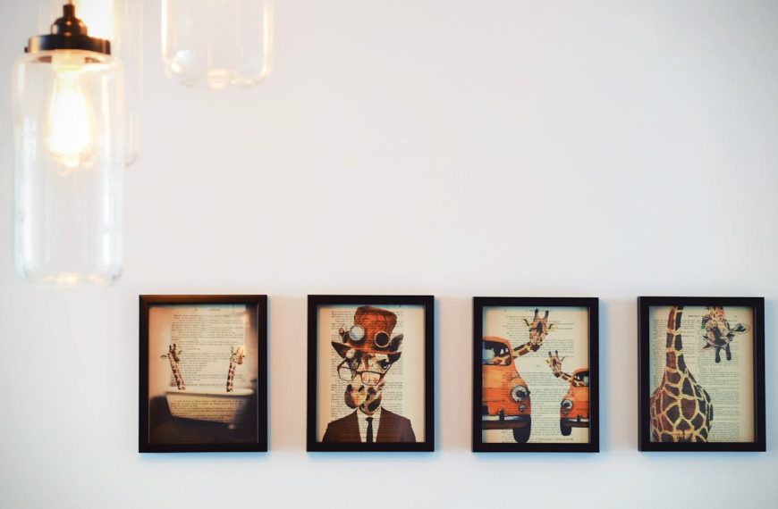 Four framed artworks featuring whimsical giraffe illustrations overlaid on vintage book pages, arranged in a row on a white wall, with hanging glass pendant lights partially visible in the upper left