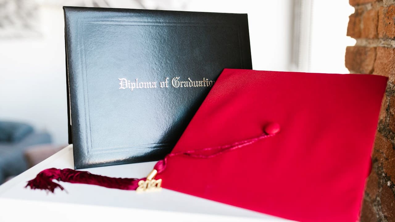 Red graduation cap with tassel, black diploma holder in the background, placed on a white surface, symbolizes academic achievement and graduation ceremony