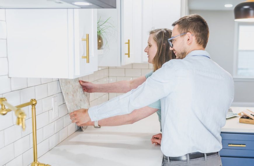 Couple holding a tile sample against a kitchen wall while discussing backsplash options