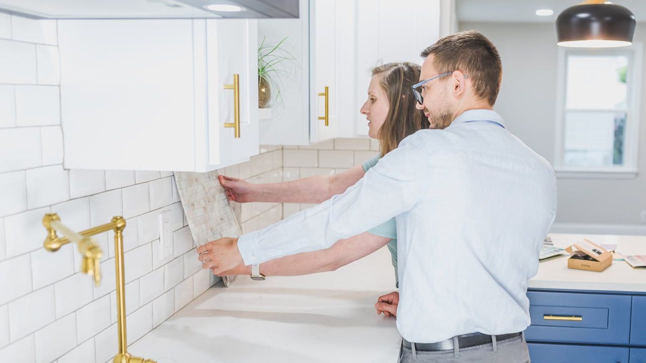Couple holding a tile sample against a kitchen wall while discussing backsplash options