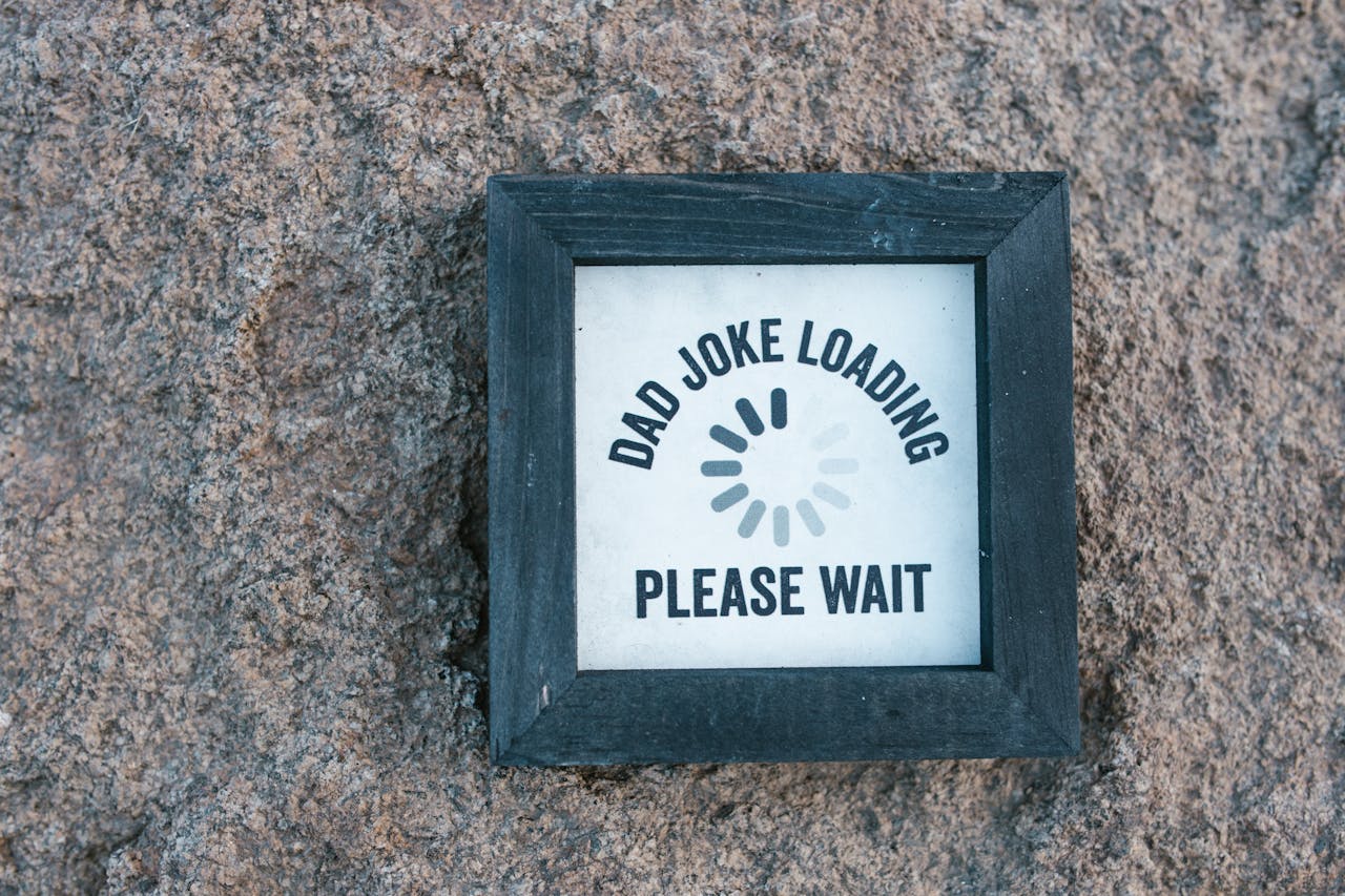 Framed sign on a stone wall, text reads "DAD JOKE LOADING" with a loading symbol beneath, followed by "PLEASE WAIT", black frame, humorous decorative plaque, playful message
