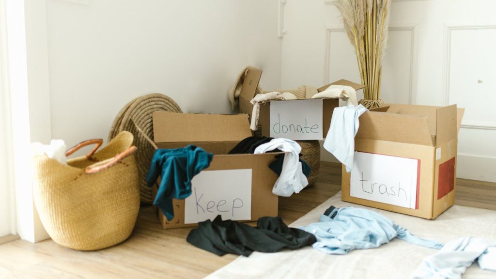 Three labeled cardboard boxes filled with clothes reading "keep", "donate", and "trash", placed in a bright room with natural light, surrounded by baskets and dried pampas grass decor