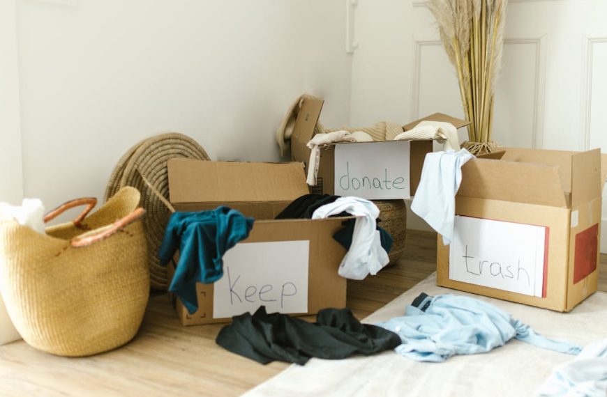Three labeled cardboard boxes filled with clothes reading "keep", "donate", and "trash", placed in a bright room with natural light, surrounded by baskets and dried pampas grass decor