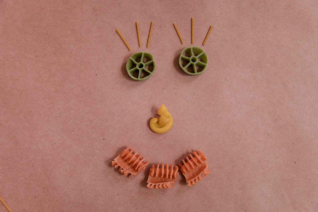 Face made from assorted pasta shapes on a brown paper background, featuring green wheel pasta eyes, spaghetti eyelashes, a yellow pasta nose, and orange pasta mouth pieces