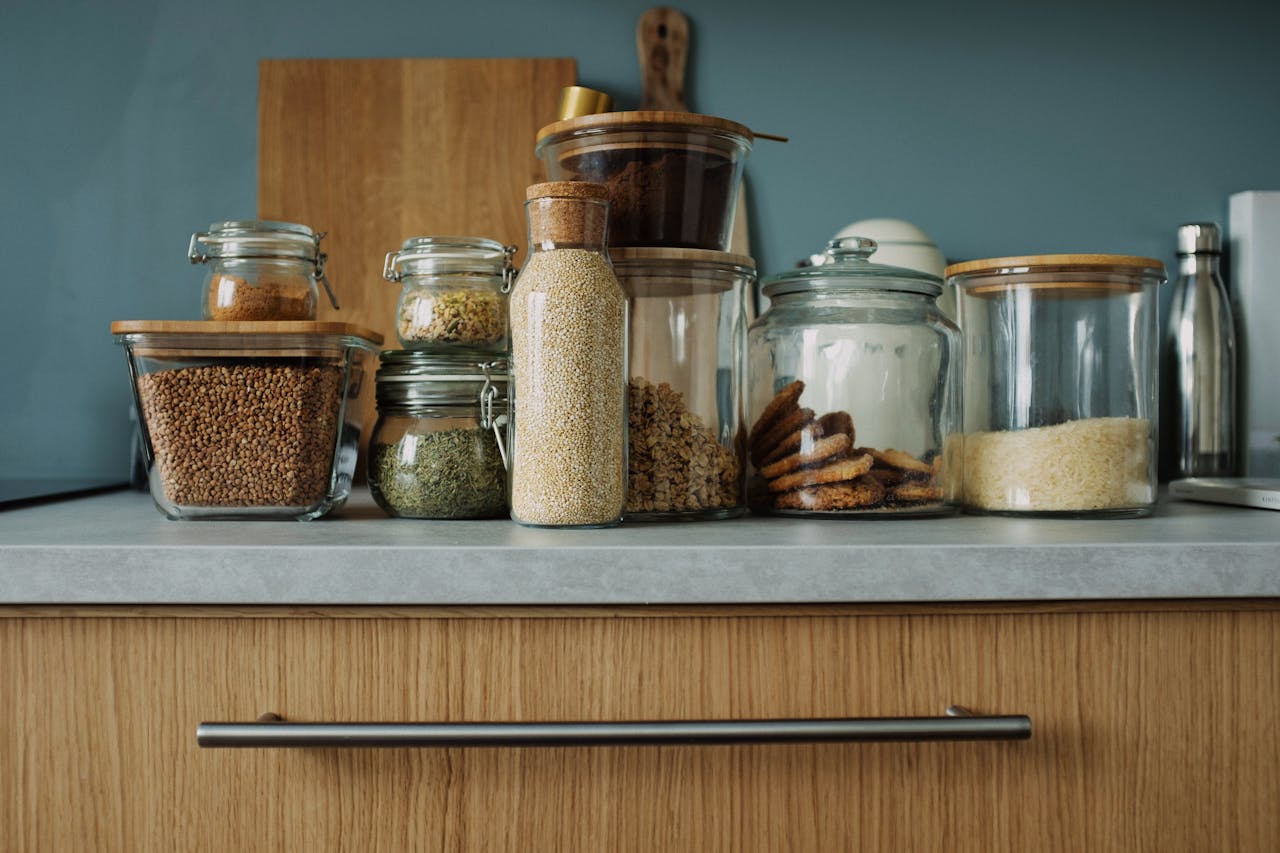Kitchen countertop with glass jars containing lentils, herbs, grains, cookies, sugar, and pasta, all neatly arranged with wooden lids, cutting board and steel bottle in the background