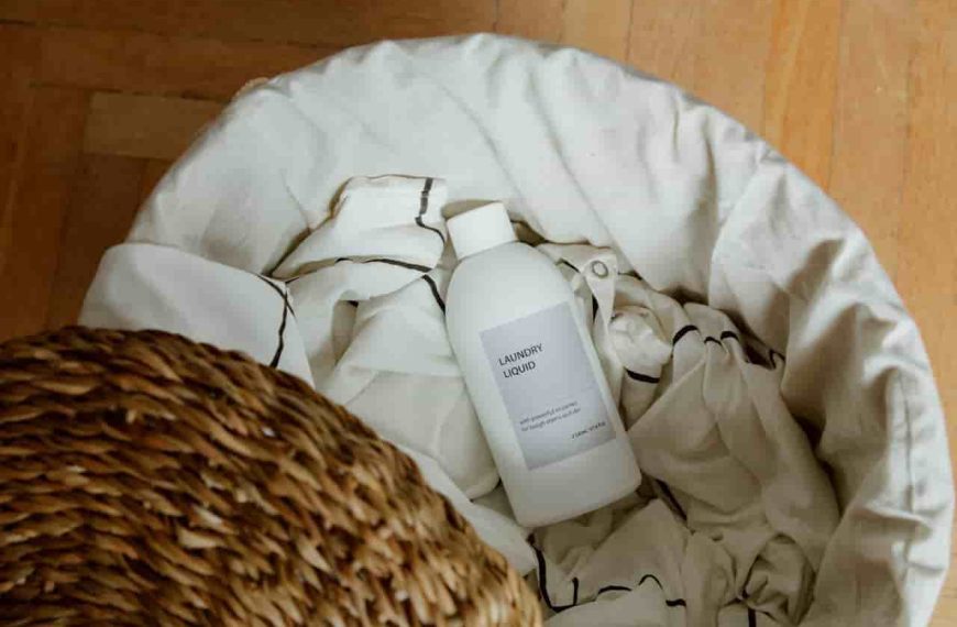 White bottle of laundry liquid, lying on light-colored fabric inside a laundry basket, partially covered with a woven wicker lid, placed on a wooden floor