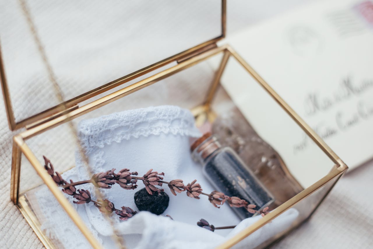 Glass box with gold metal edges, containing a white lace-trimmed cloth, dried lavender sprig, small black stone, and a corked glass vial with dark contents