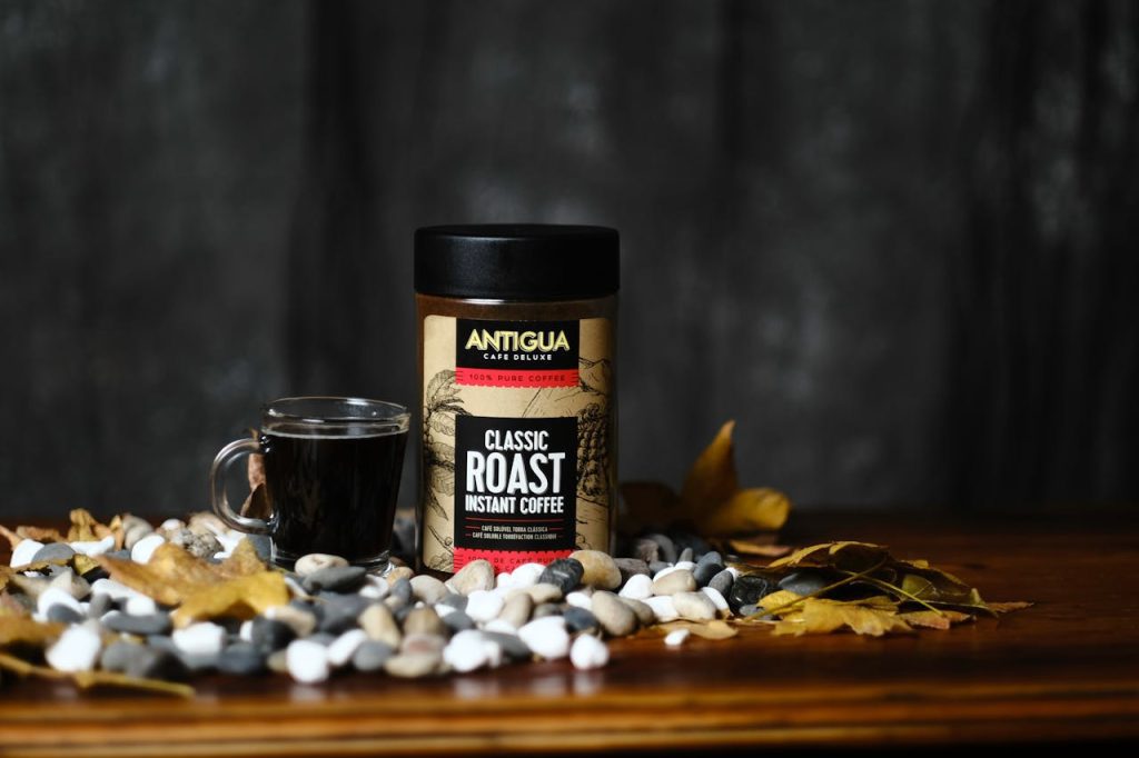 Jar of Nescafé Gold instant coffee, placed on a wooden surface, surrounded by coffee beans, a clear glass cup of black coffee, decorative leaves, and a dark textured background