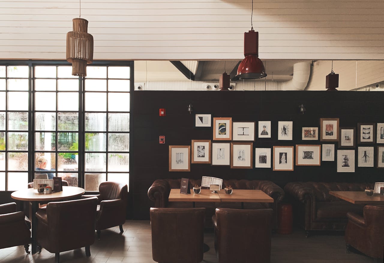 Cozy café interior with brown leather armchairs, wooden tables, dark paneled wall decorated with a grid of framed minimalist art, large grid-style window letting in natural light