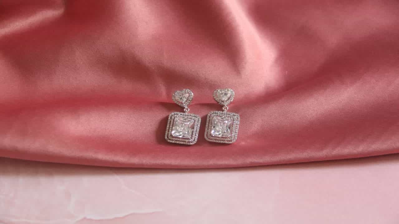 Pair of sparkly silver earrings with square-cut gemstones and heart-shaped tops, displayed on soft pink satin fabric