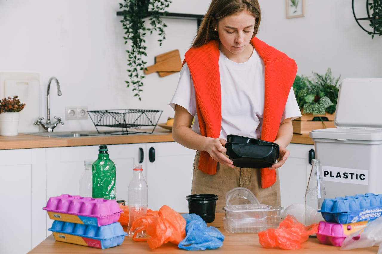 Woman sorting plastic containers and bottles, standing in a kitchen, surrounded by plastic waste, next to a labeled plastic recycling bin
