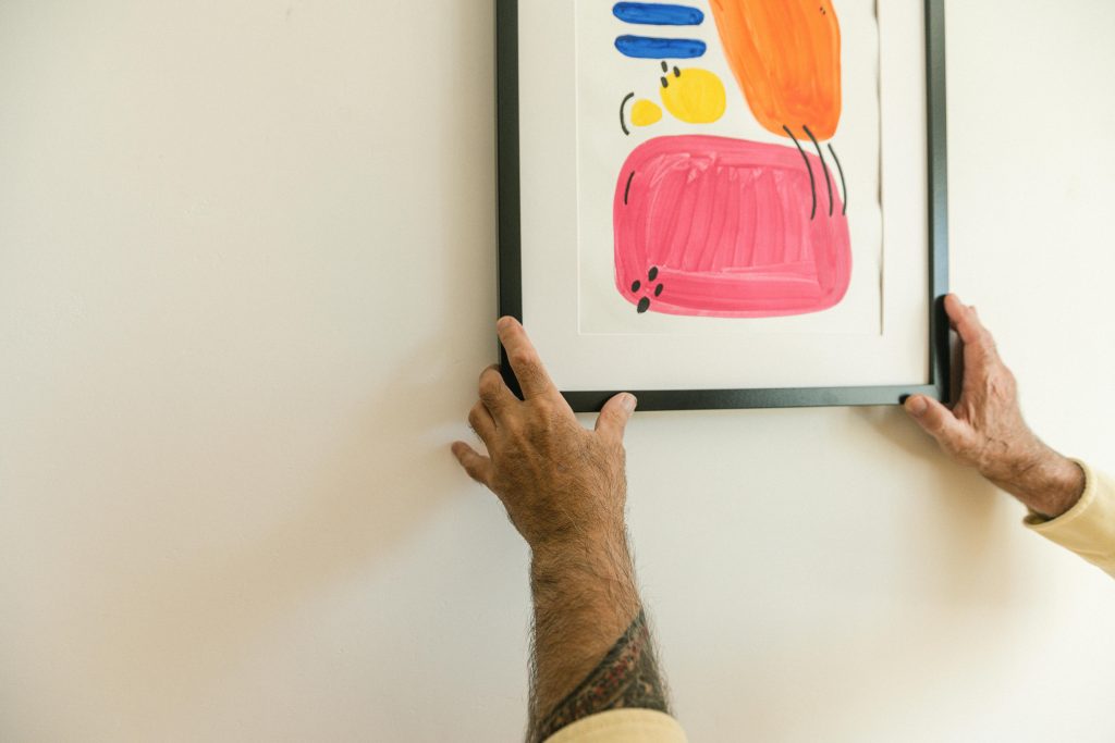 Man’s tattooed arms adjusting a colorful abstract painting in a black frame on a light wall