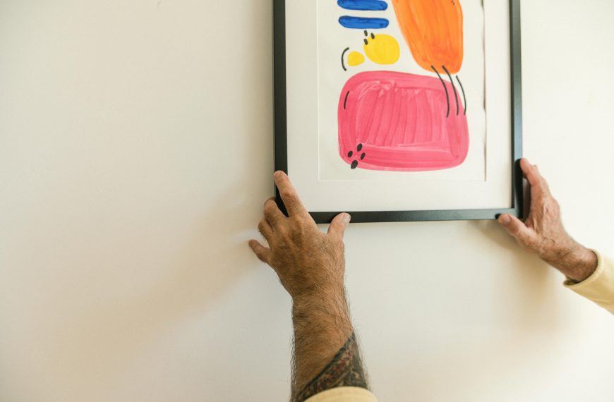 Man’s tattooed arms adjusting a colorful abstract painting in a black frame on a light wall