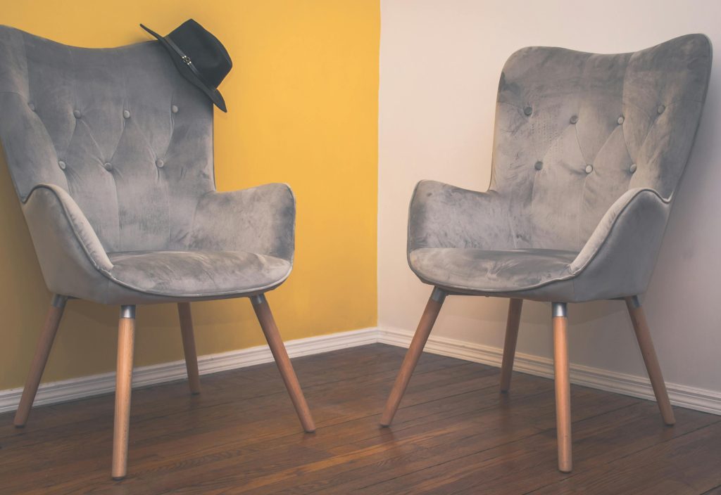 Two modern grey upholstered chairs with wooden legs, placed in the corner of a room with wooden flooring, one chair has a black hat resting on its backrest, yellow and white painted walls in the background