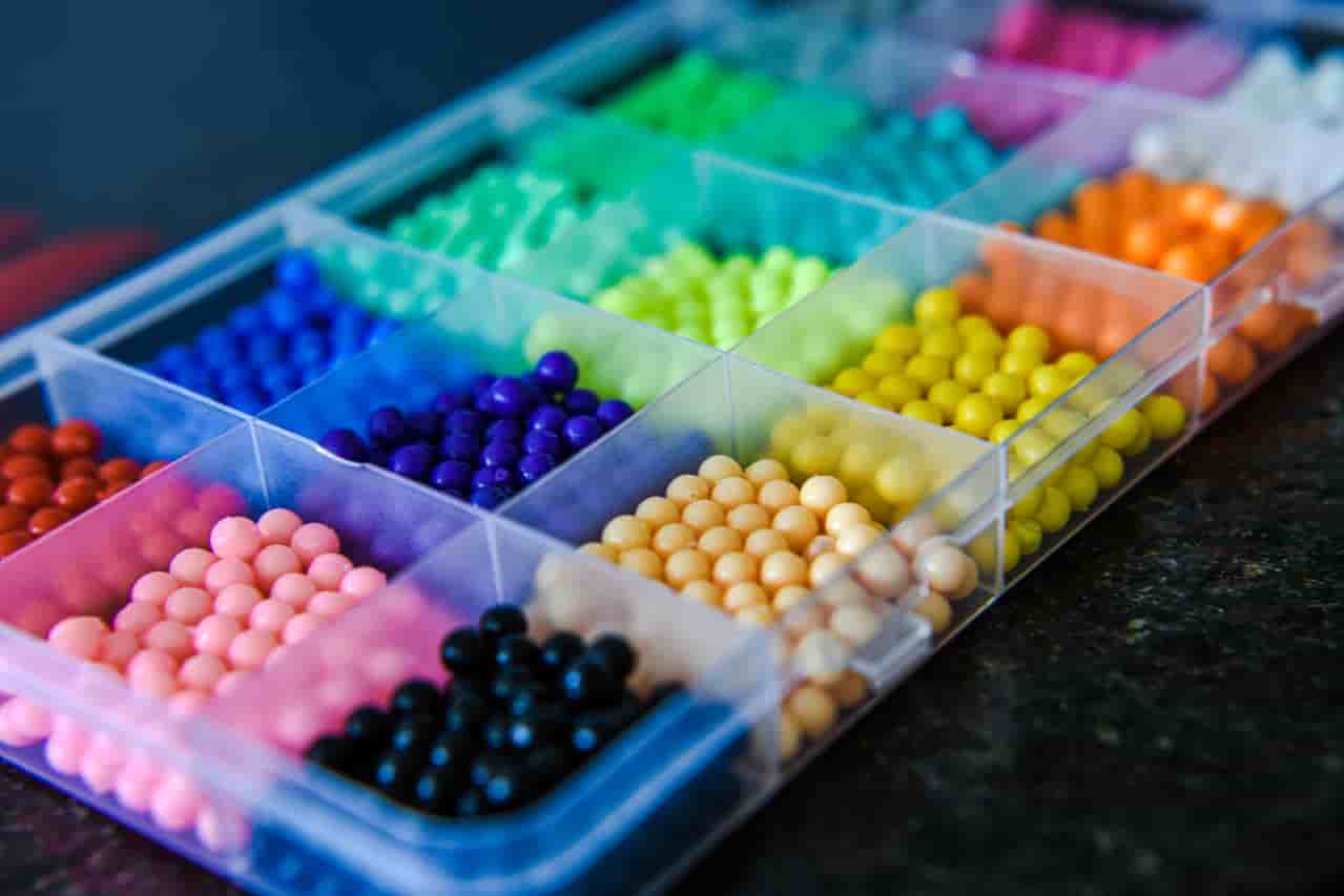 Colorful round beads arranged in a clear plastic organizer, multiple compartments, beads in shades of pink, black, blue, green, yellow, and orange