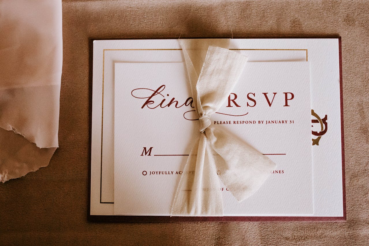 Elegant RSVP card with gold border, burgundy script font, and a cream fabric ribbon, placed on a soft beige surface beside a translucent fabric