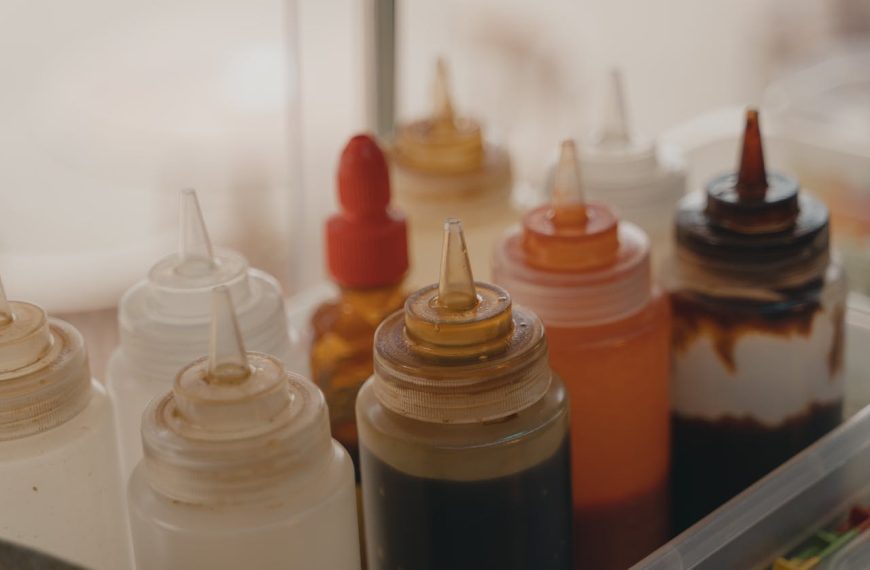 Close-up of assorted plastic squeeze bottles filled with various sauces and condiments, including white, brown, red, and dark mixtures, stored together in a clear plastic container