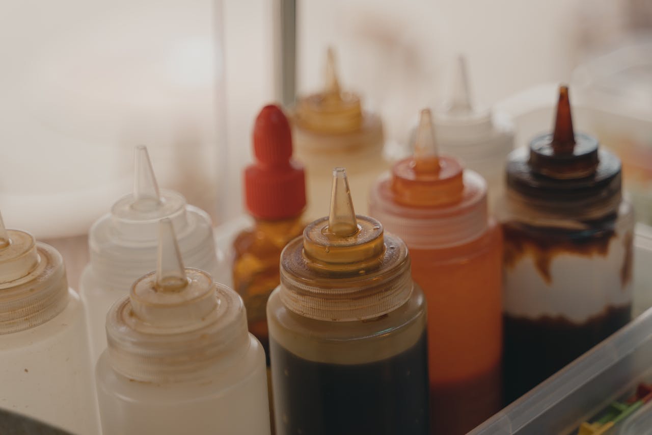 Close-up of assorted plastic squeeze bottles filled with various sauces and condiments, including white, brown, red, and dark mixtures, stored together in a clear plastic container