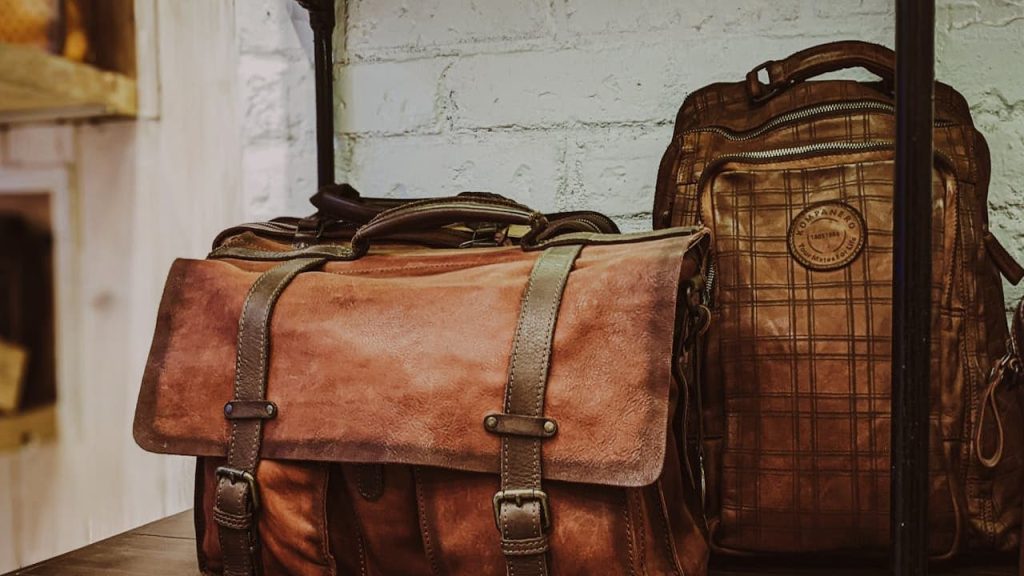 Two vintage leather bags on a dark wooden shelf, front bag is a satchel with buckle straps and multiple pockets, back bag is a backpack with a plaid pattern and rounded top, both in brown tones, set against a white brick wall