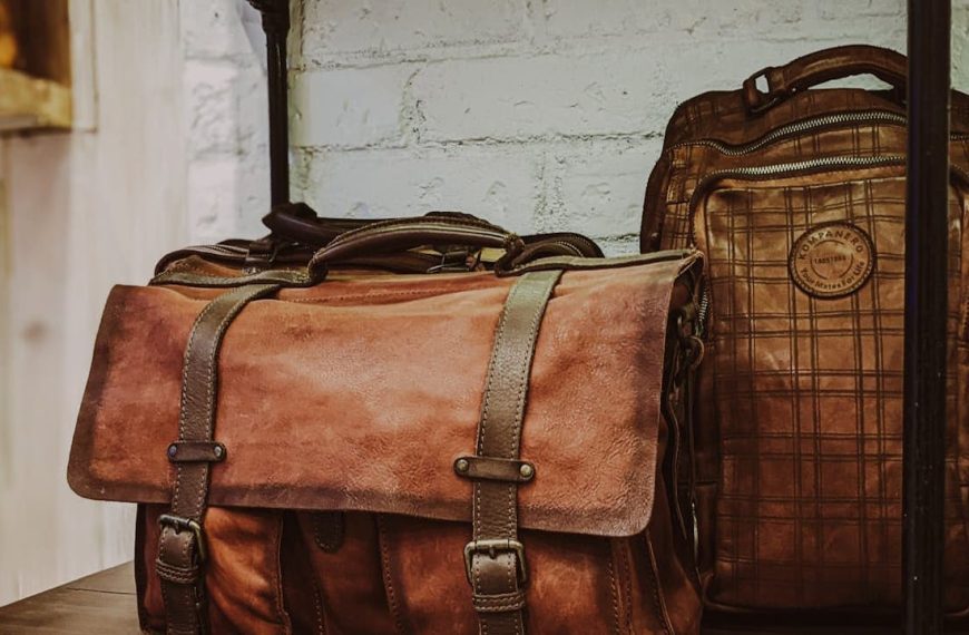 Two vintage leather bags on a dark wooden shelf, front bag is a satchel with buckle straps and multiple pockets, back bag is a backpack with a plaid pattern and rounded top, both in brown tones, set against a white brick wall