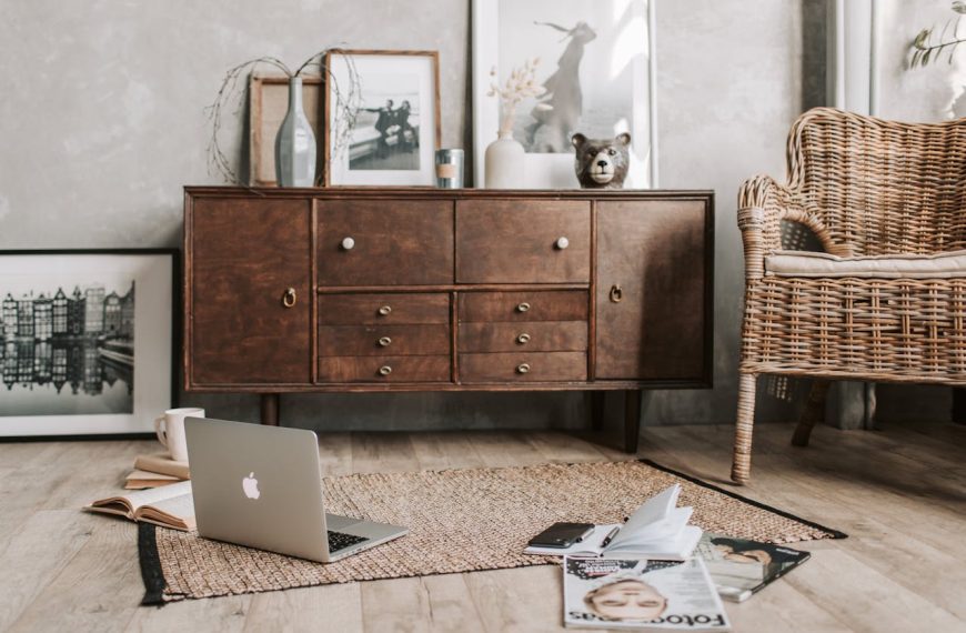Cozy living room corner with a wooden sideboard, framed black-and-white photos, a rattan armchair, woven rug, open laptop, books, coffee mug, magazines