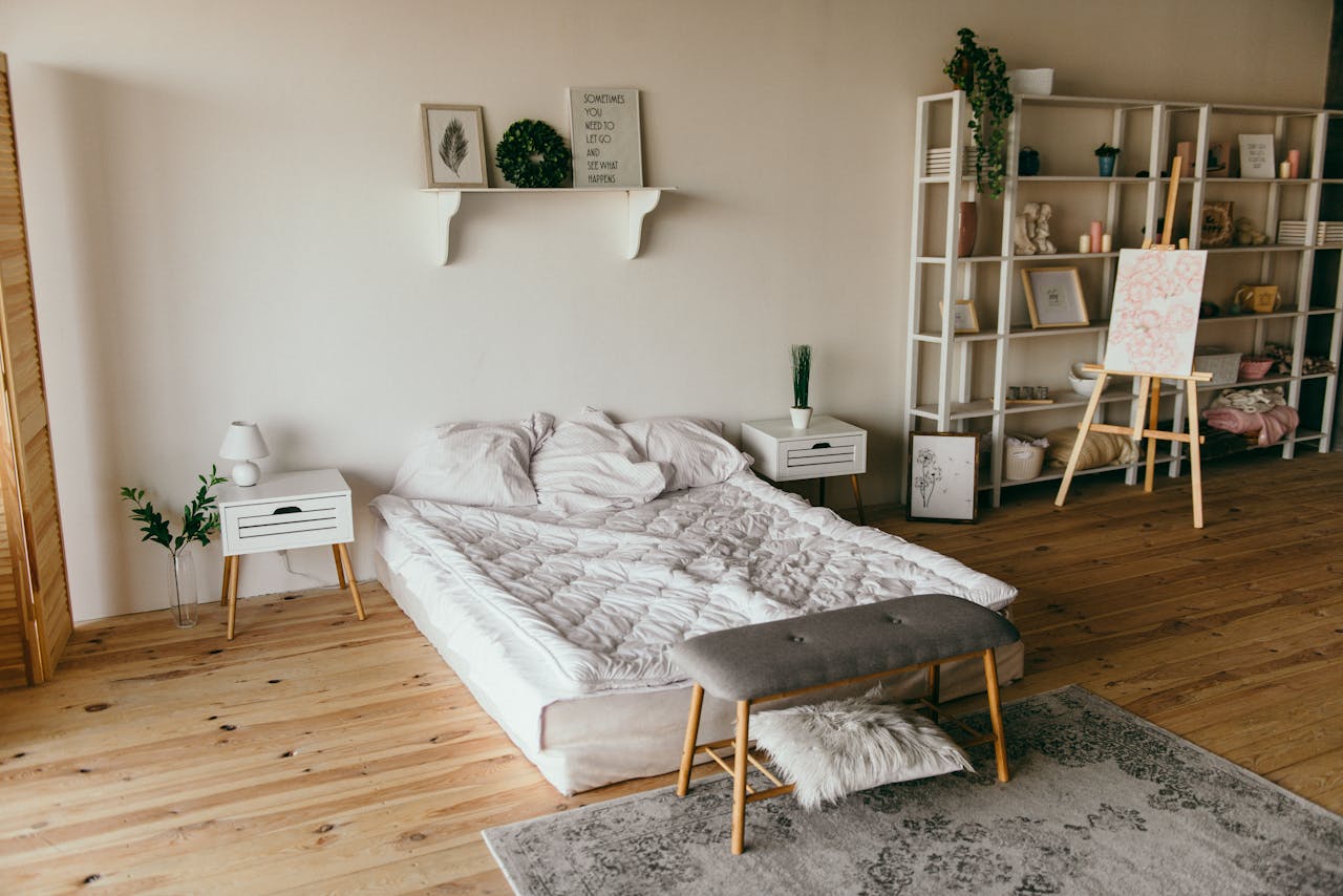 Minimalist bedroom with a low bed, white bedding, two white nightstands with lamps and plants, wooden floor, a bench at the foot of the bed, and a white shelf filled with books, candles, and art supplies