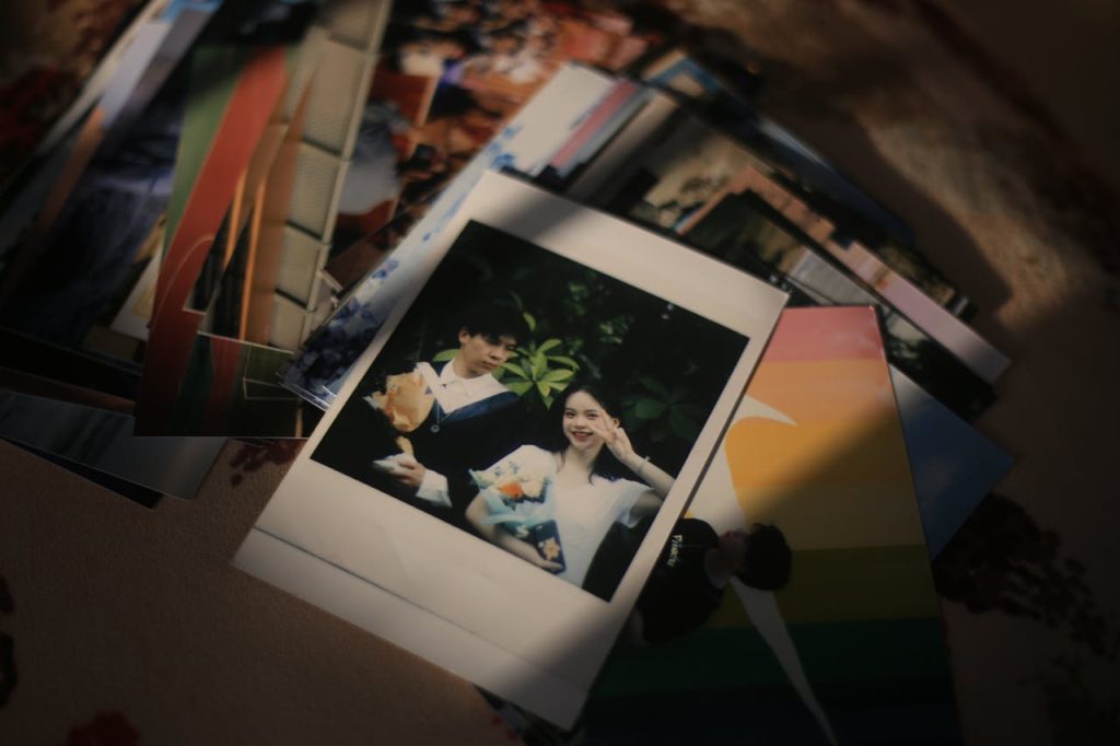 A pile of printed photographs scattered on a surface, with a prominent Polaroid showing two smiling people holding flower bouquets