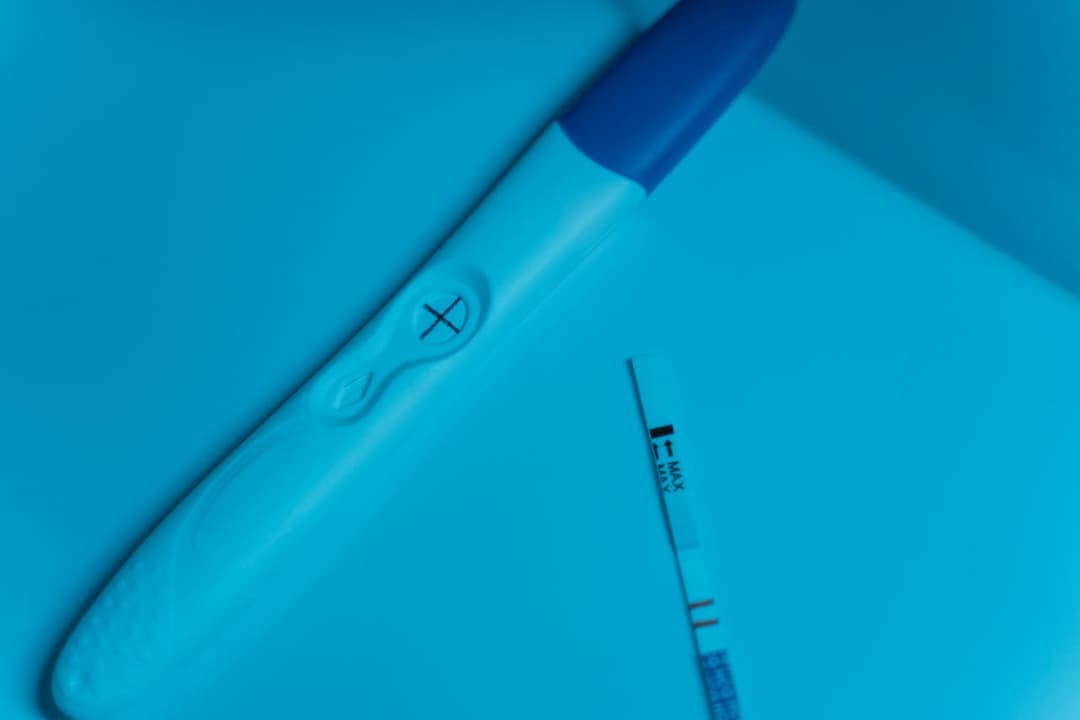Positive pregnancy test with blue cap and clear cross symbol, placed on a blue surface beside a pregnancy test strip showing two lines