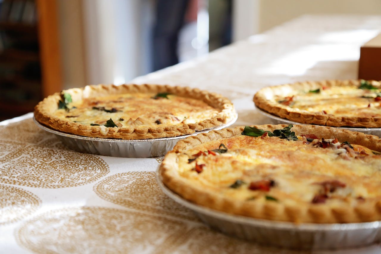 Three freshly baked quiches in aluminum pie tins, with golden, flaky crusts and vibrant toppings, placed on a decorative, patterned tablecloth in natural sunlight