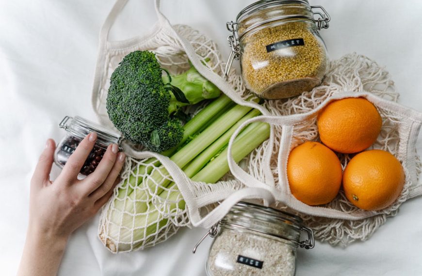 Reusable mesh bags filled with fresh vegetables and fruits including broccoli, celery, corn, and oranges, jars of grains and peanut butter