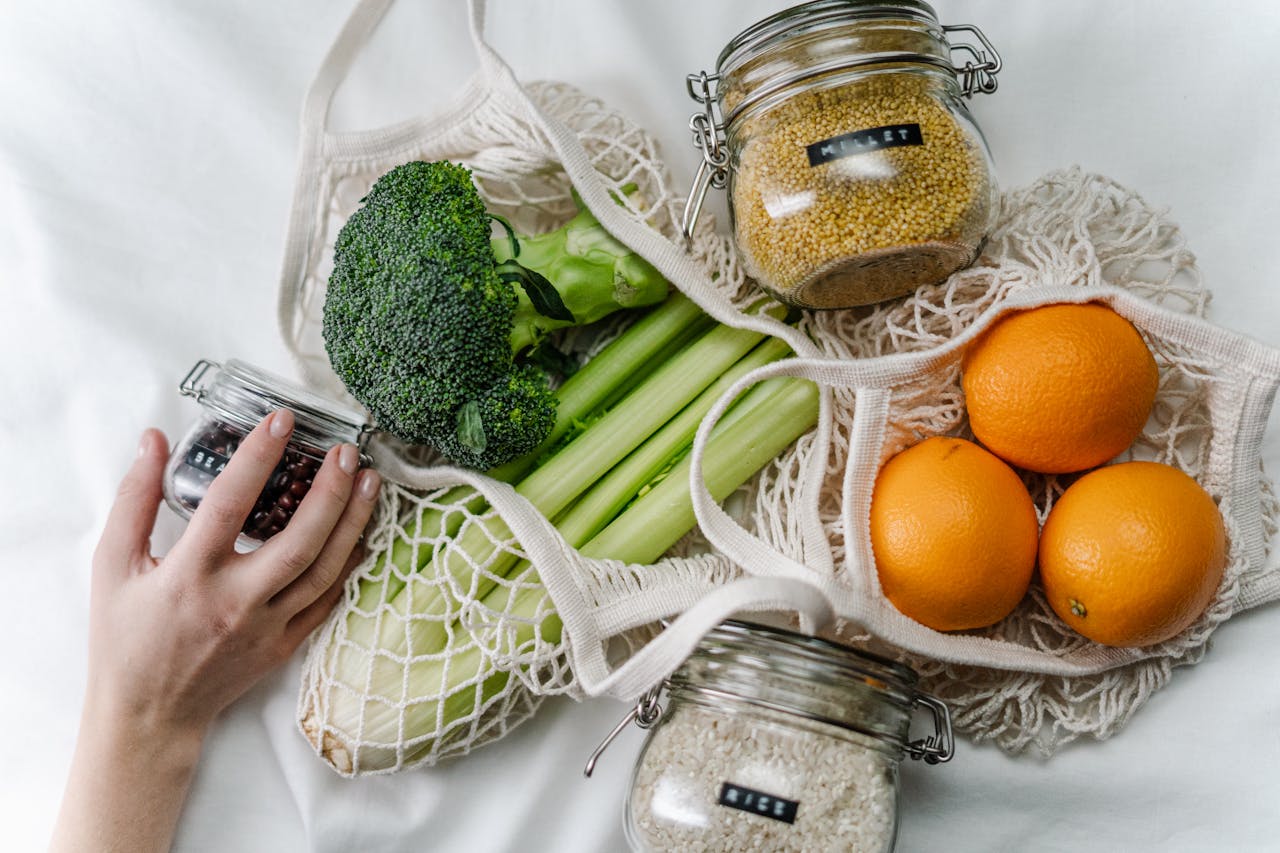 Reusable mesh bags filled with fresh vegetables and fruits including broccoli, celery, corn, and oranges, jars of grains and peanut butter