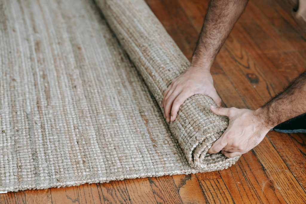 Hands rolling up a textured beige jute rug on a polished wooden floor, close-up view