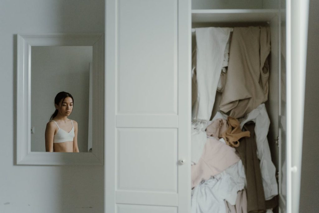 Young woman standing solemnly in front of a mirror, reflected next to a messy, overstuffed wardrobe filled with disorganized clothes in a dimly lit room