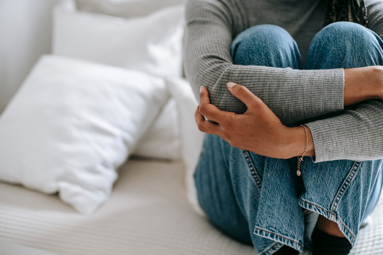 Person sitting on a bed hugging their knees, wearing a grey sweater and blue jeans, surrounded by white pillows, body language suggesting sadness or emotional distress