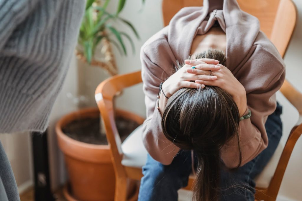 Woman sitting in a chair with her head down and hands clasped over her head, body tense, suggesting emotional distress or overwhelm