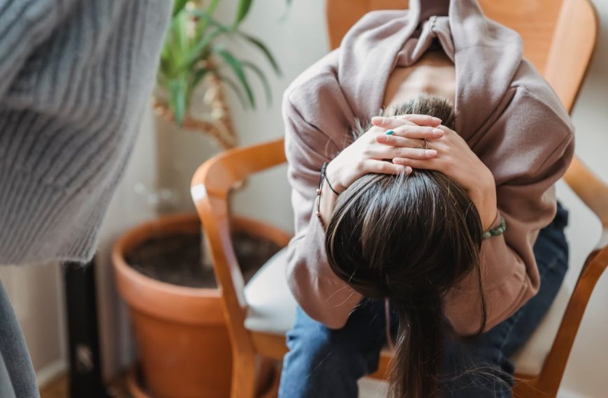 Woman sitting in a chair with her head down and hands clasped over her head, body tense, suggesting emotional distress or overwhelm