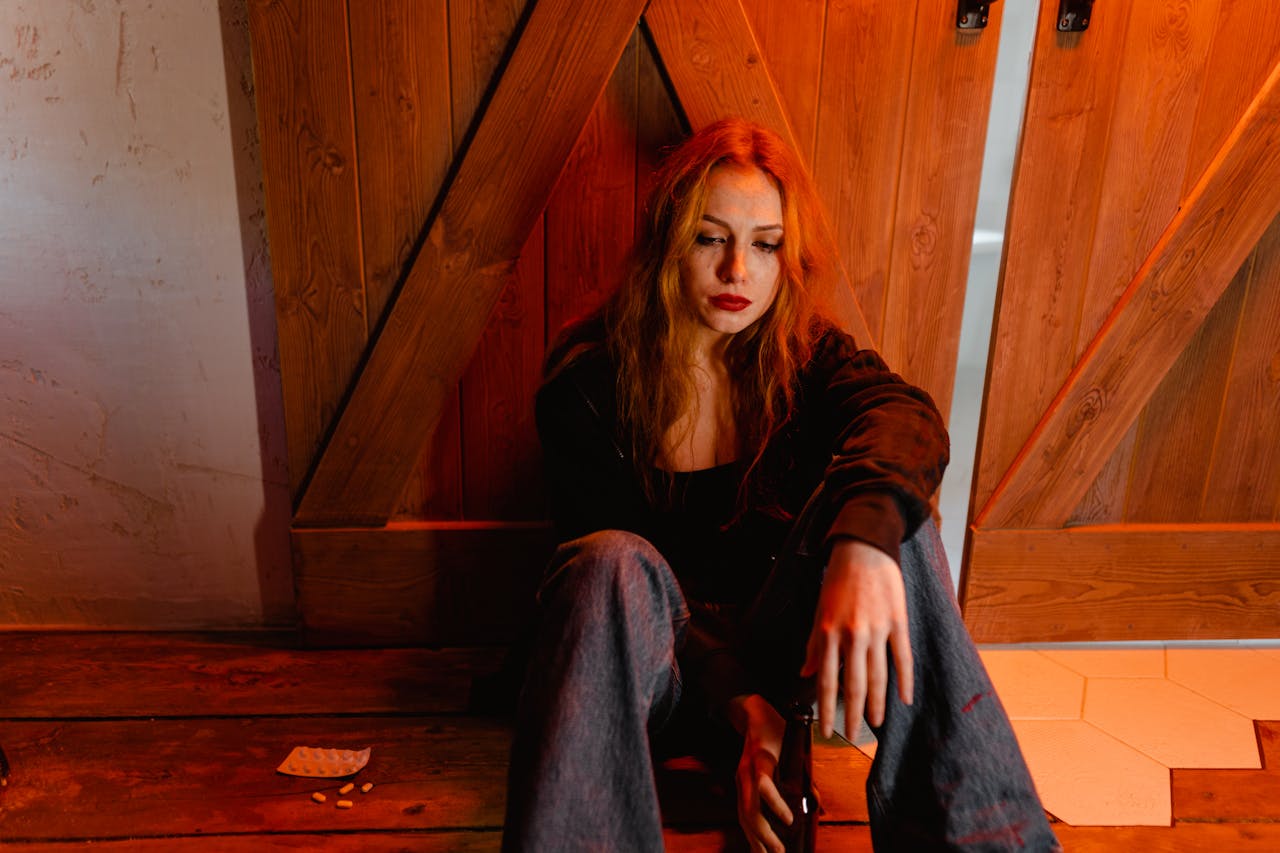 Sad woman with smeared makeup sitting on a wooden floor against wooden doors, holding a bottle, with pills scattered nearby, under red lighting