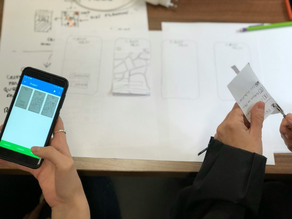 Hands holding a smartphone and a small paper document, scanning papers on a desk, multiple documents laid out, smartphone screen showing a scanning app, digitizing physical papers