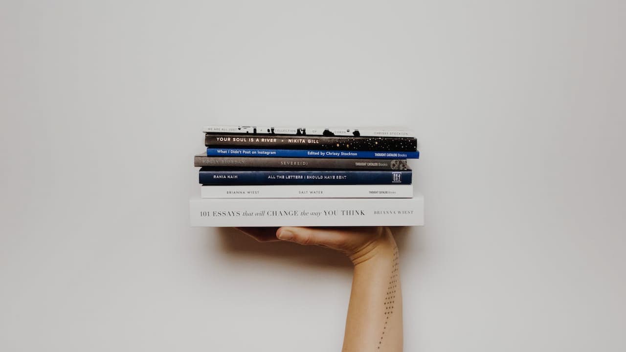 Stack of self-help and poetry books balanced on an outstretched hand with tattooed forearm, against a plain white background