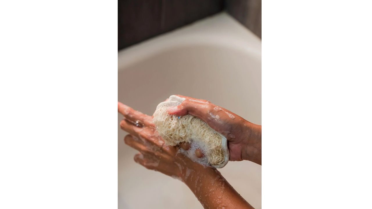 Person washing with a natural loofah sponge in a soapy lather, hands covered in suds, bathtub in background