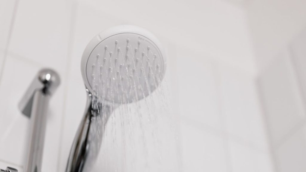 Chrome handheld shower head releasing a steady stream of water, mounted on a wall with white ceramic tiles, close-up view
