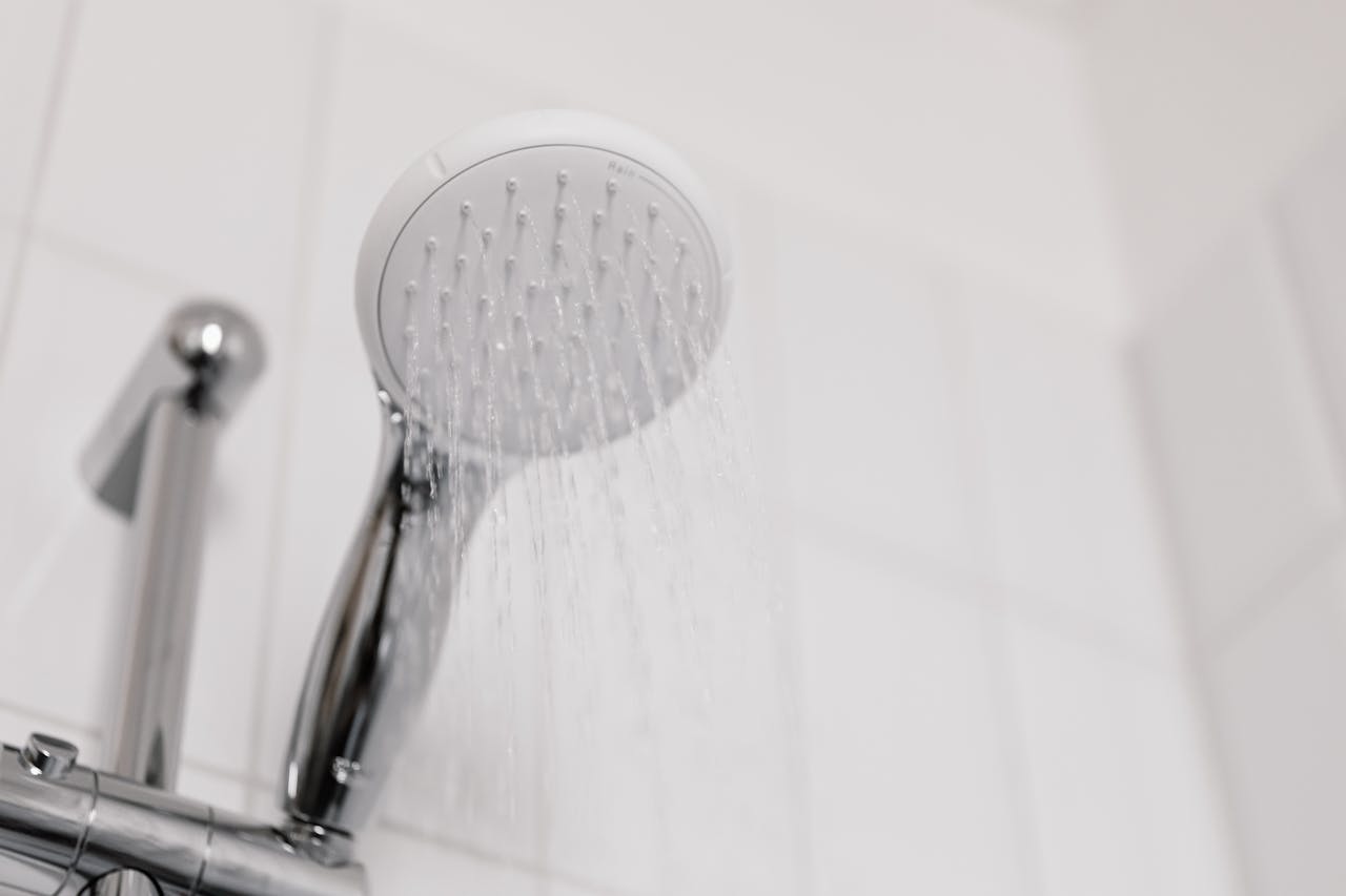 Chrome handheld shower head releasing a steady stream of water, mounted on a wall with white ceramic tiles, close-up view