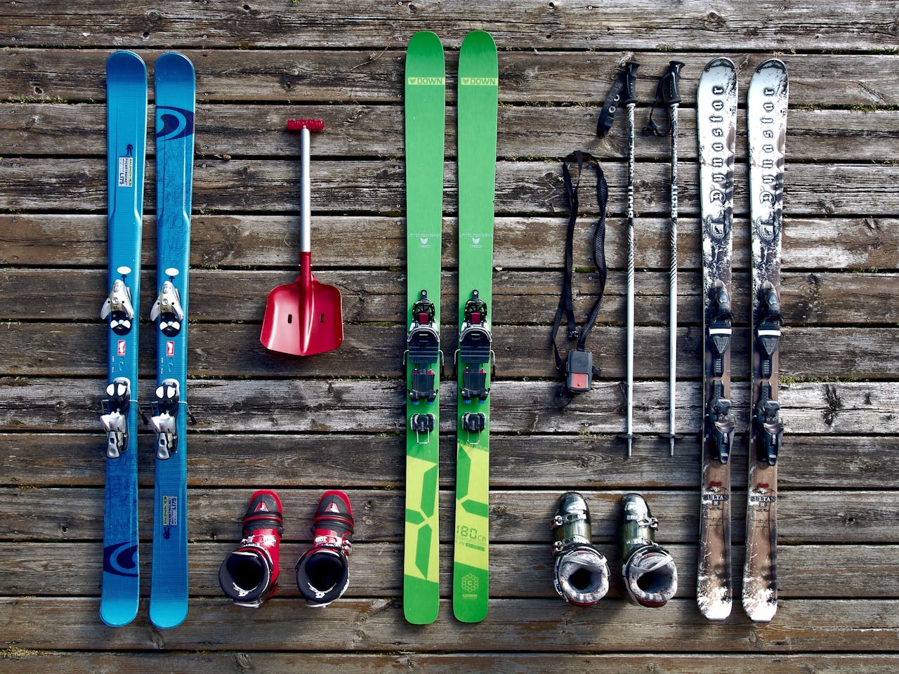 Three pairs of skis, two sets of ski boots, ski poles, avalanche beacon, and a red snow shovel neatly arranged on a weathered wooden deck