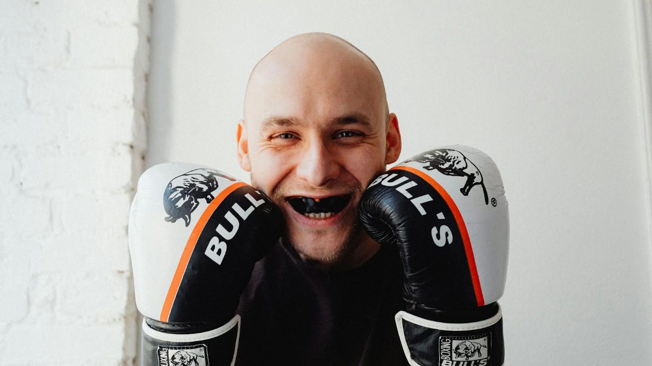 Smiling man wearing boxing gloves and a black mouthguard, posing playfully in a boxing stance against a white background