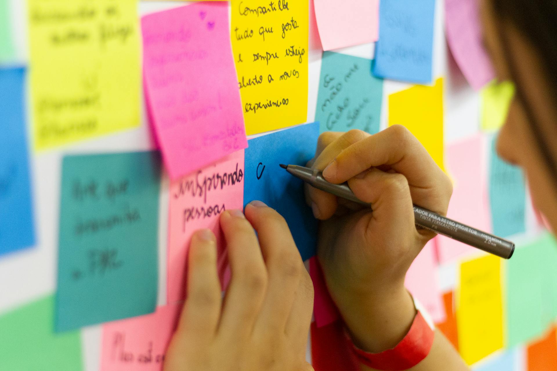 Hand writing on a blue sticky note, surrounded by multiple colorful sticky notes on a wall, notes contain handwritten tasks or reminders, close-up view, vibrant and organized workspace