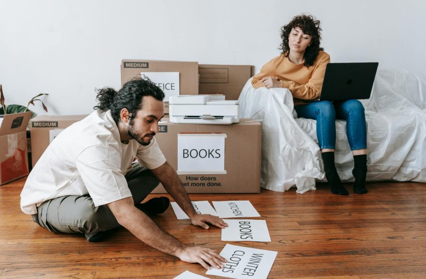 Two people unpacking moving boxes in empty room with hardwood floors, one kneeling, one sitting on box