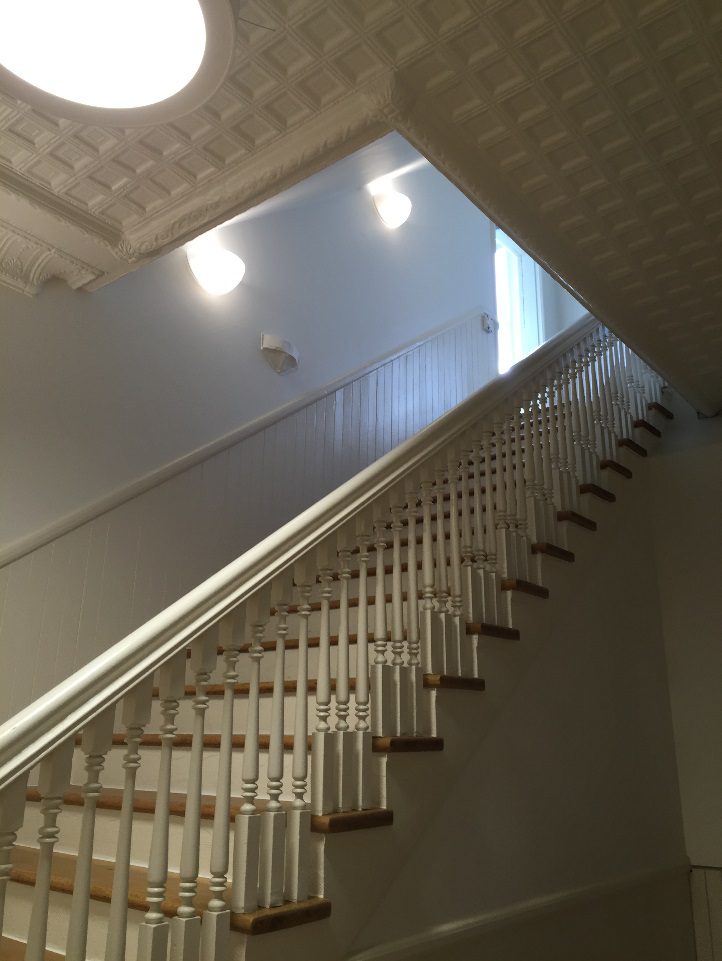 Indoor staircase with white railings, wooden steps, ceiling with patterned tiles, lit by recessed ceiling lights, natural light entering from a window at the top of the stairs
