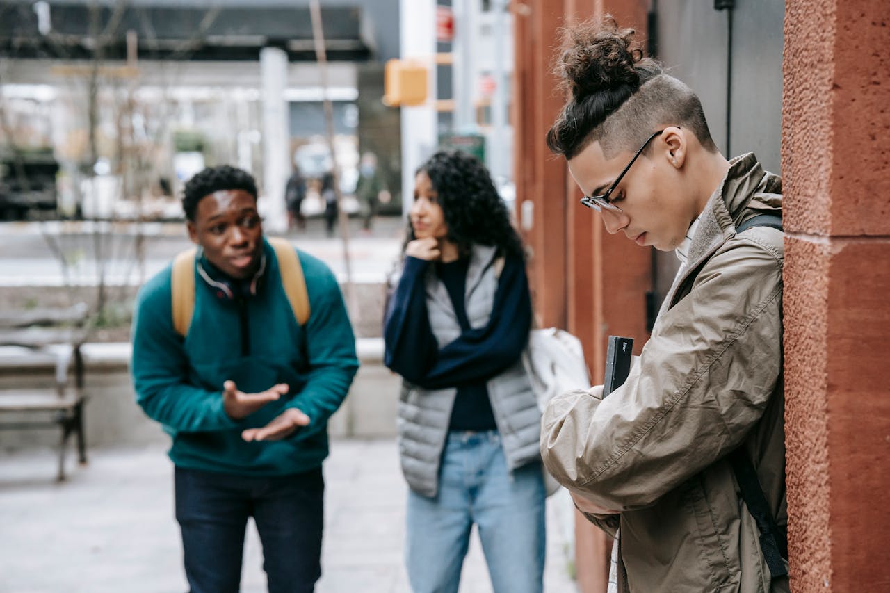 Three young adults standing outside, one person leaning against a wall looking down while the other two appear to be talking intensely, possibly arguing or confronting