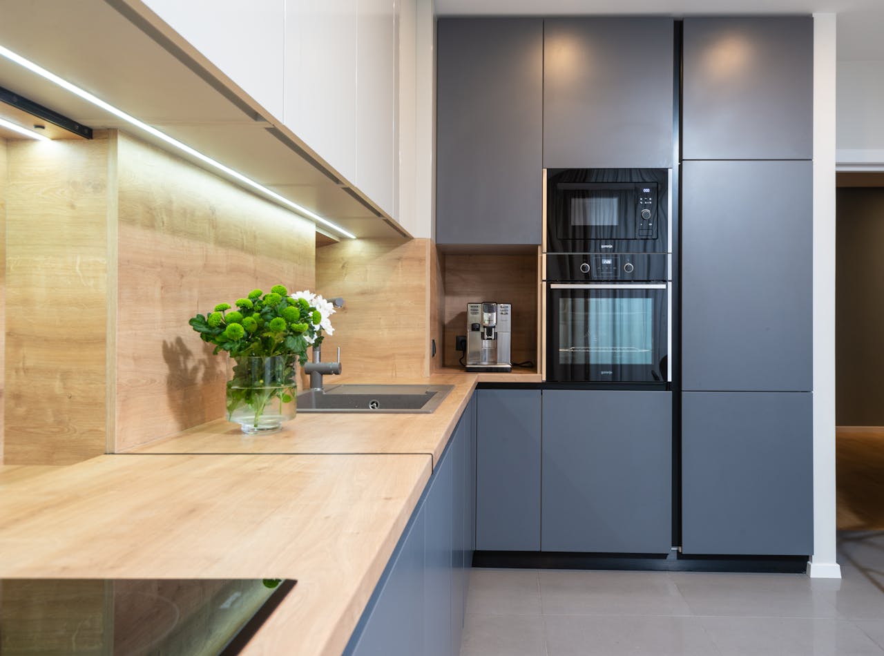 Modern kitchen interior, sleek grey cabinets, built-in oven and microwave, wooden countertops and backsplash, under-cabinet lighting, potted green plant on counter, minimalist design